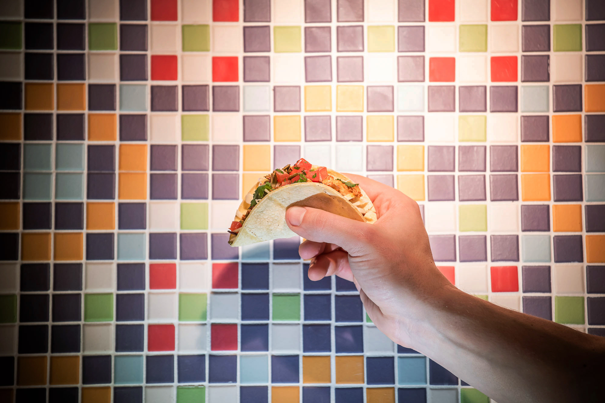 hand holds taco in front of colourful tile